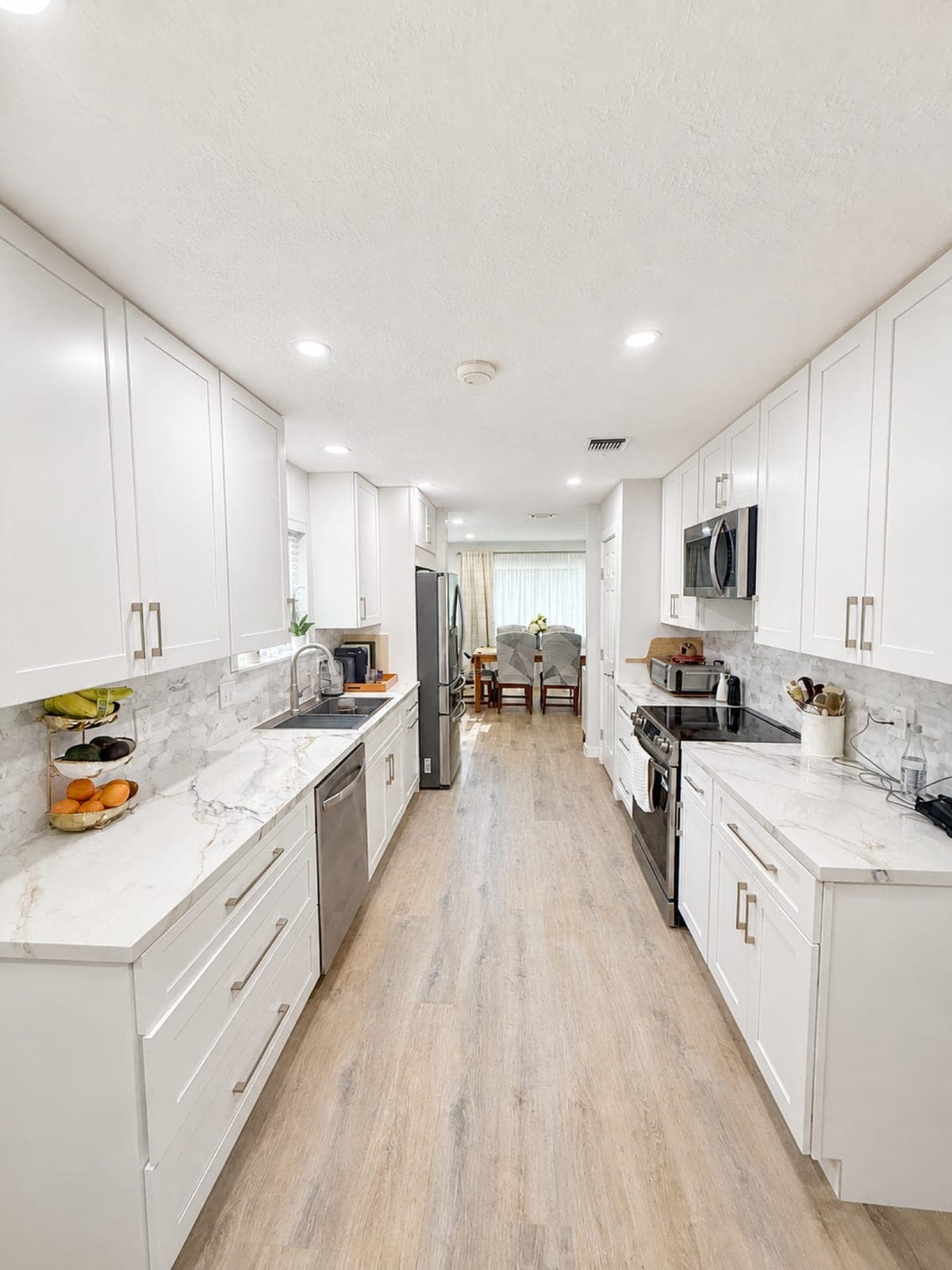 The finished galley kitchen: white shaker cabinets, marbled quartz counters and backsplash, stainless appliances, and pale wood-look flooring running through to the dining room