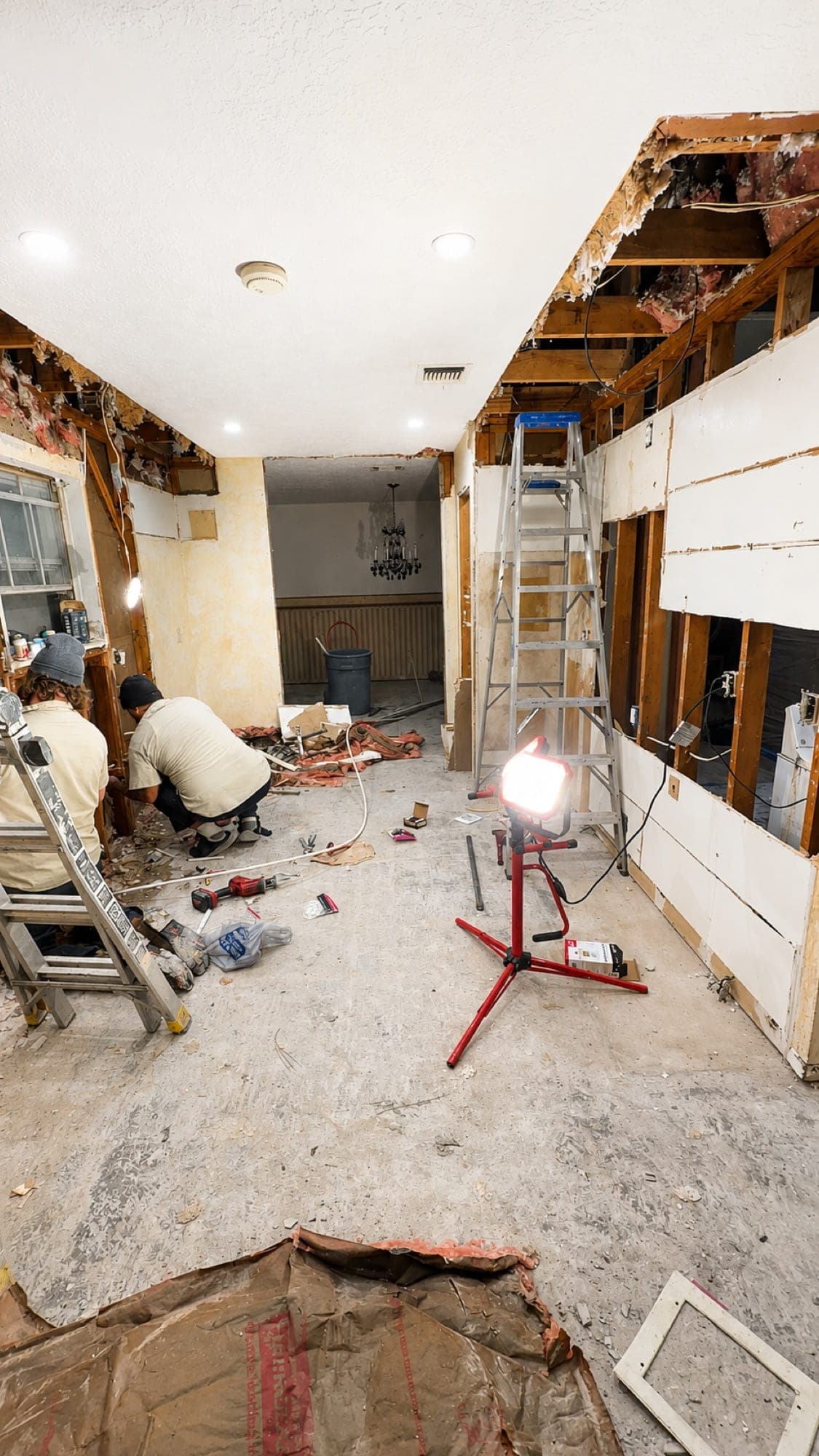 The same kitchen mid-renovation: walls stripped to the studs, a work light and ladder on a bare concrete floor, and two crew members working along the left wall