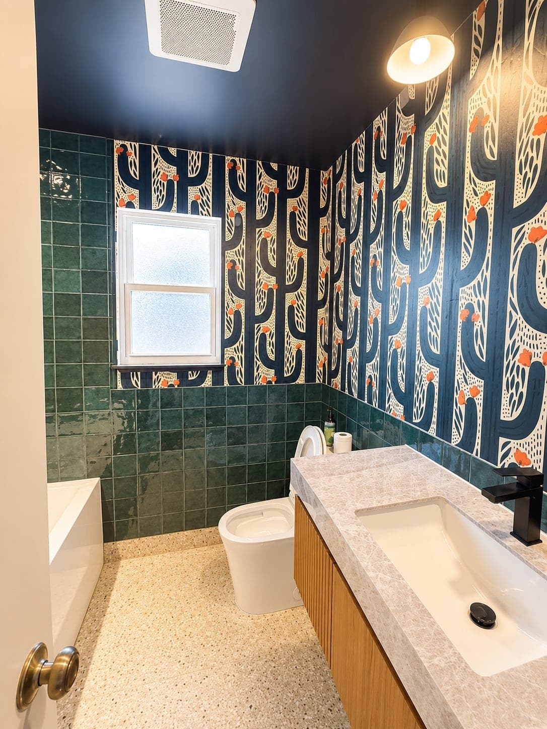 The finished bathroom: bold navy cactus wallpaper, glossy green tile, a floating stone vanity with an integrated sink and black faucet, and warm terrazzo flooring