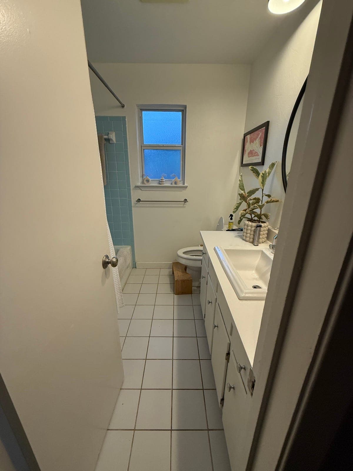 The same bathroom before: plain white wall and floor tile, a basic vanity with a large mirror, and a frosted window